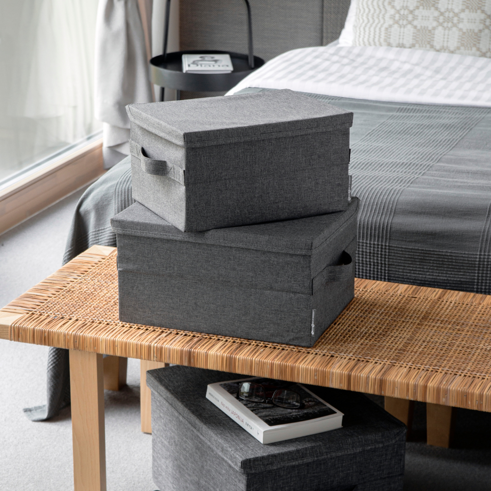 Twee grijze SOFT-opbergboxen (45x34x25 cm) van Bigso staan opgestapeld op een gevlochten bankje aan het voeteneinde van een bed. Ernaast liggen een kleinere box en een boek, allemaal zacht verlicht door het natuurlijke licht dat door het raam naar binnen valt.