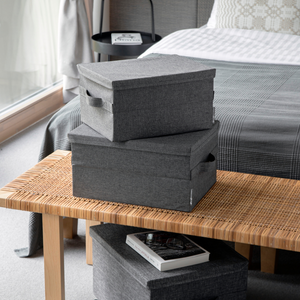 Twee grijze SOFT-opbergboxen (45x34x25 cm) van Bigso staan opgestapeld op een gevlochten bankje aan het voeteneinde van een bed. Ernaast liggen een kleinere box en een boek, allemaal zacht verlicht door het natuurlijke licht dat door het raam naar binnen valt.