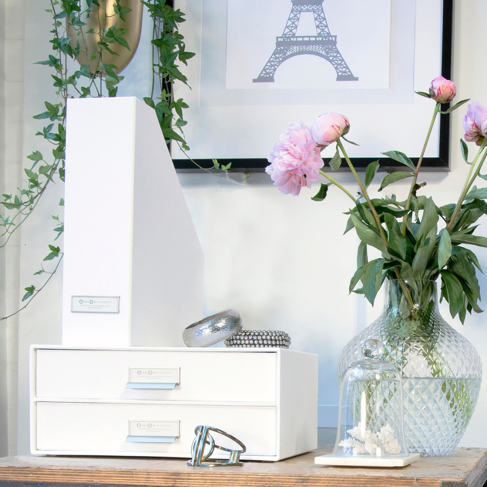 Een opgeruimd bureau met witte lades, de Bigso VIKTORIA staande tijdschriftenhouder – wit met etikettenhouder, een glazen vaas met roze pioenrozen, een glazen stolp met witte koraal, groene ranken en een ingelijste afbeelding van de Eiffeltoren aan de muur.