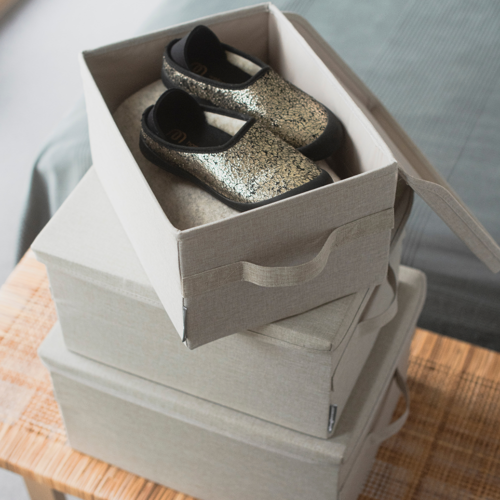 Een paar zwart-gouden loopschoentjes zit in een open, opvouwbare doos op twee gesloten, stapelbare SOFT-opbergdozen Beige L - 45x34x25 cm van Bigso, op een houten oppervlak naast een bed.