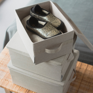 Een paar zwart-gouden loopschoentjes zit in een open, opvouwbare doos op twee gesloten, stapelbare SOFT-opbergdozen Beige L - 45x34x25 cm van Bigso, op een houten oppervlak naast een bed.