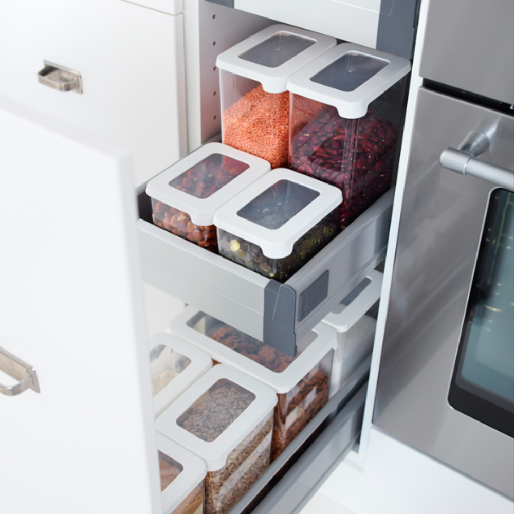 Een Orthex SmartStore-set met doorzichtige en witte voorraadpotten, stuk voor stuk luchtdicht afgesloten en gevuld met granen, linzen, bonen en pasta, staat overzichtelijk gerangschikt op uittrekbare planken in een keukenkast naast de oven.