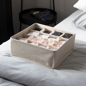 De SOFT Organizer Beige – 16 vakken van Bigso, gemaakt van stof, biedt de perfecte opbergruimte voor kleding in verschillende kleuren. Hij staat op een bed met lichtgrijs beddengoed, met daarnaast een zwart bijzettafeltje en lavendelkleurige decoratie.