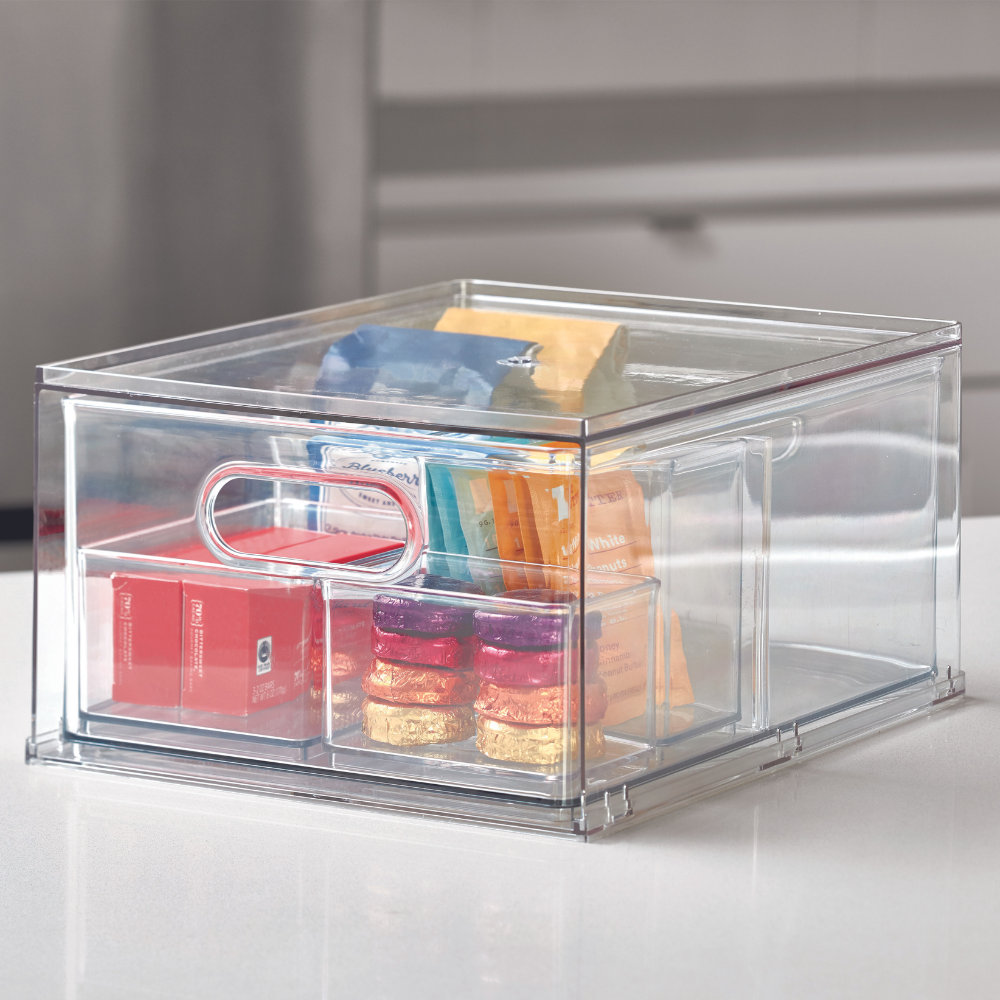 Een doorzichtige iDesign HOME EDIT-opbergdoos met handvat (35 x 26,7 x 19,7 cm), gevuld met allerlei kleurrijke snacks, staat op een wit aanrechtblad in een moderne keuken.