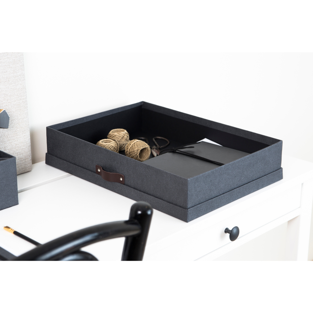 Een SVERKER opbergbox A3 in zwart canvas van Bigso staat op een wit bureau, waarop garenklossen, scharen en notitieboekjes netjes zijn opgeborgen. Een zwarte stoel en een potlood maken het minimalistische, overzichtelijke tafereel compleet.