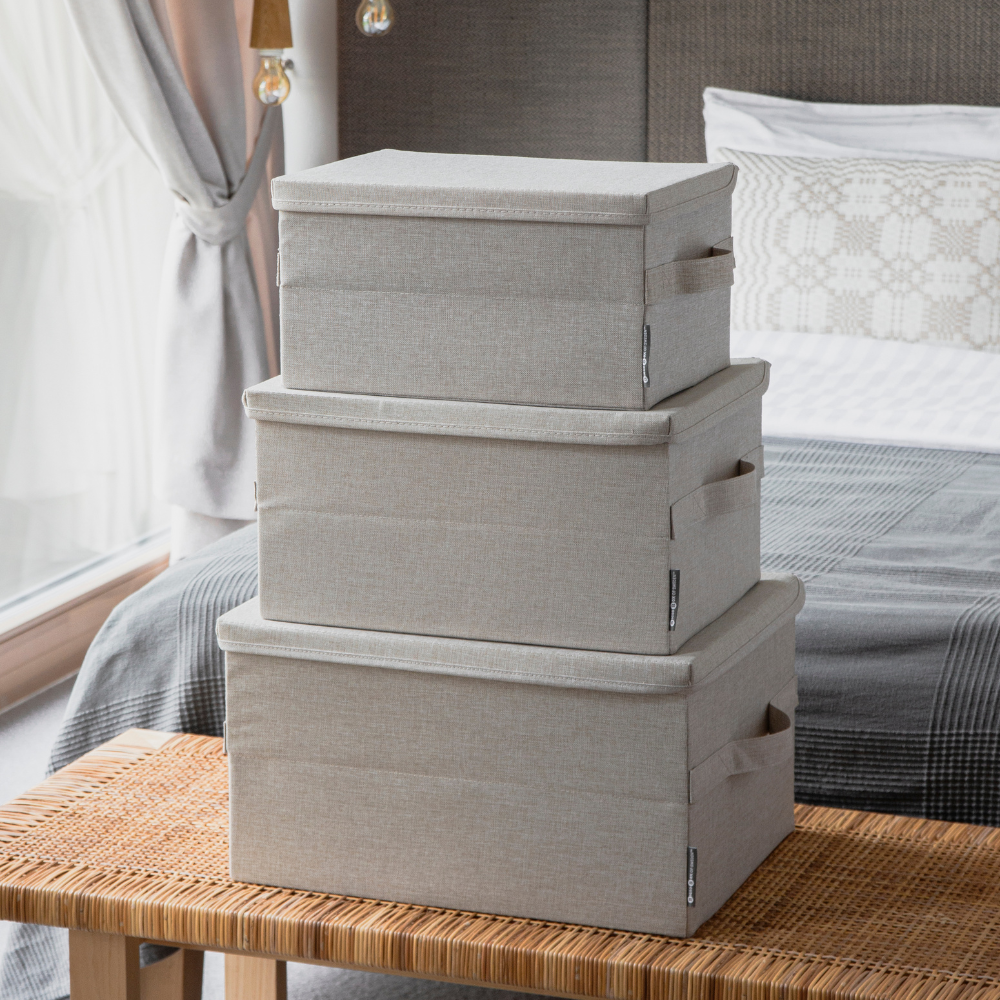 Drie Bigso SOFT opbergboxen in beige (maat M, 40x30x22 cm) staan op een bank in de slaapkamer opgestapeld; op de achtergrond zie je een net opgemaakt bed en een raam met transparante gordijnen.