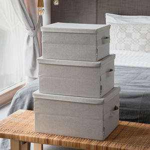 Drie Bigso SOFT opbergboxen in beige (maat M, 40x30x22 cm) staan op een bank in de slaapkamer opgestapeld; op de achtergrond zie je een net opgemaakt bed en een raam met transparante gordijnen.