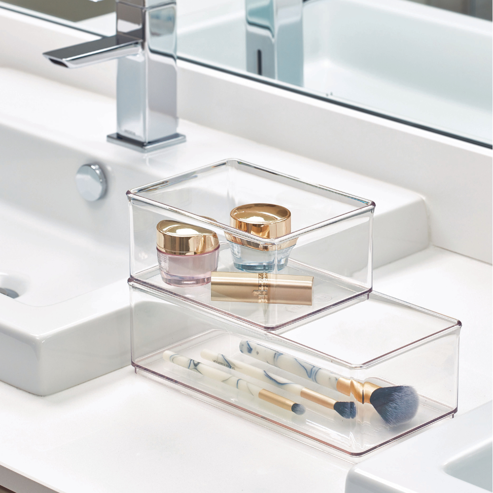 Doorzichtige opbergdozen met cosmeticaproducten en kwasten op een witte wastafel naast de kraan en de spiegel.