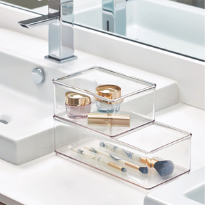 Doorzichtige opbergdozen met cosmeticaproducten en kwasten op een witte wastafel naast de kraan en de spiegel.