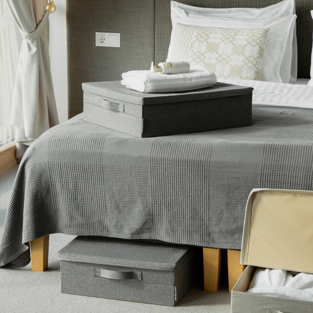 Een netjes opgemaakt bed met een grijs, gestructureerd sprei en kussens is voorzien van een SOFT opbergbox Grijs – opbergruimte onder het bed van Bigso voor slimme opbergruimte onder het bed, terwijl er een open koffer aan het voeteneinde van het bed staat.