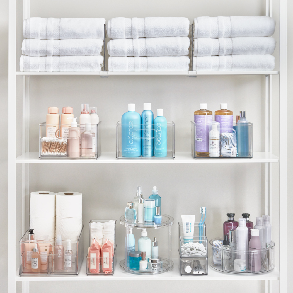 Een witte plank biedt ruimte voor opgestapelde handdoeken, iDesign HOME EDIT opbergbakjes KLAR (25,4 x 25,4 x 15,2 cm), rollen toiletpapier en doorzichtige bakjes met gesorteerde hygiëne- en schoonheidsproducten voor een opgeruimde, stapelbare opbergoplossing.