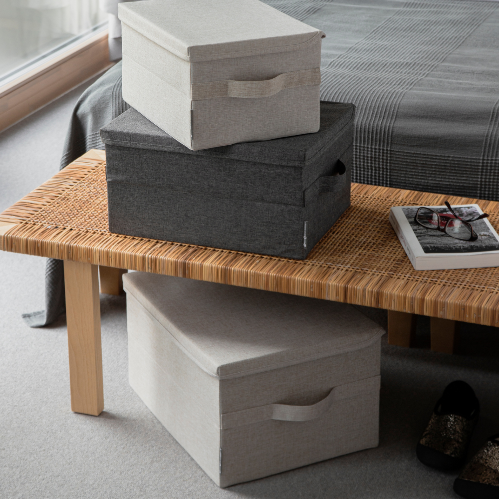Drie Bigso SOFT opbergboxen Beige L (45x34x25 cm): twee staan op een gevlochten houten bank gestapeld, één staat eronder. Ernaast liggen een tijdschrift, een bril en een afstandsbediening. Op de achtergrond staat een bed met een geruite deken.