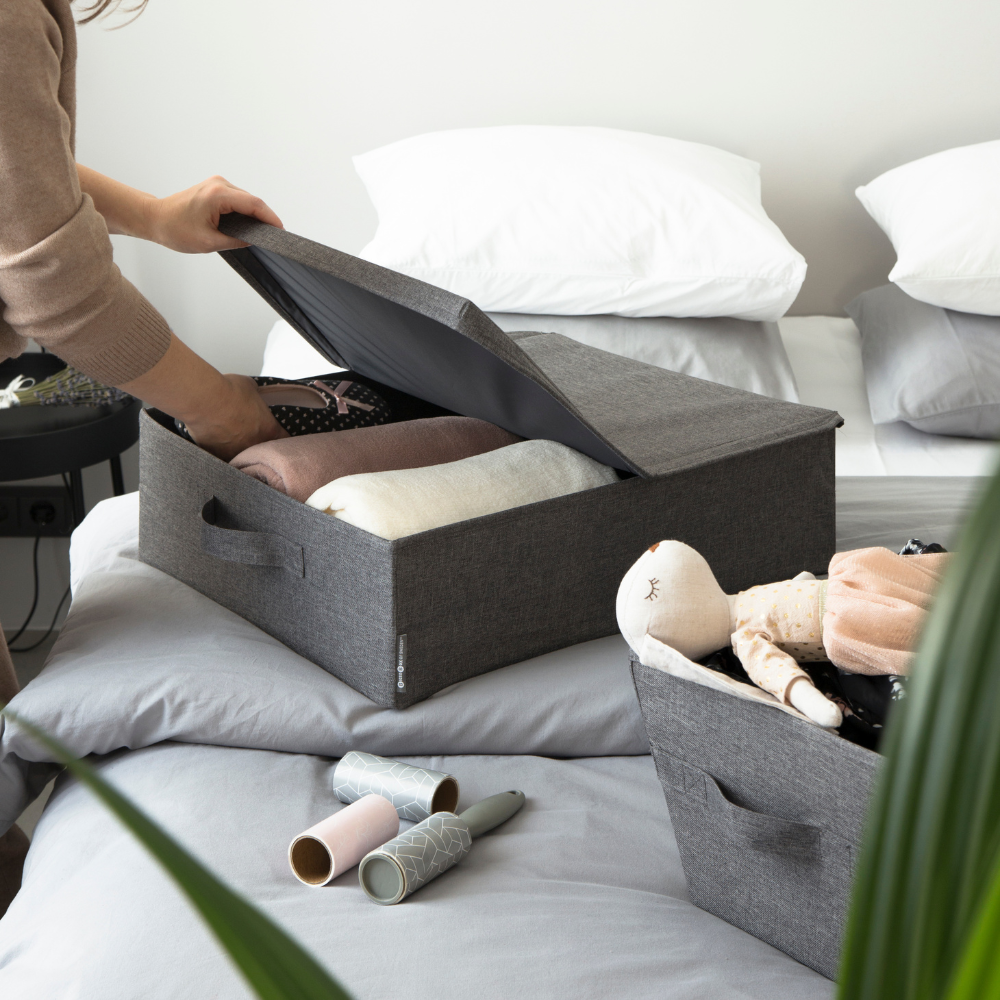 Iemand legt kleding op in de Bigso SOFT opbergbox Grijs – opbergruimte onder het bed – op een opgeruimd bed met witte kussens. Er zijn een knuffel, pluisrollers en een groene plant te zien, die de stijlvolle opbergoplossing van de box nog eens extra benadrukken.