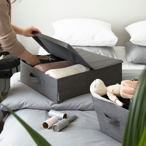 Iemand legt kleding op in de Bigso SOFT opbergbox Grijs – opbergruimte onder het bed – op een opgeruimd bed met witte kussens. Er zijn een knuffel, pluisrollers en een groene plant te zien, die de stijlvolle opbergoplossing van de box nog eens extra benadrukken.