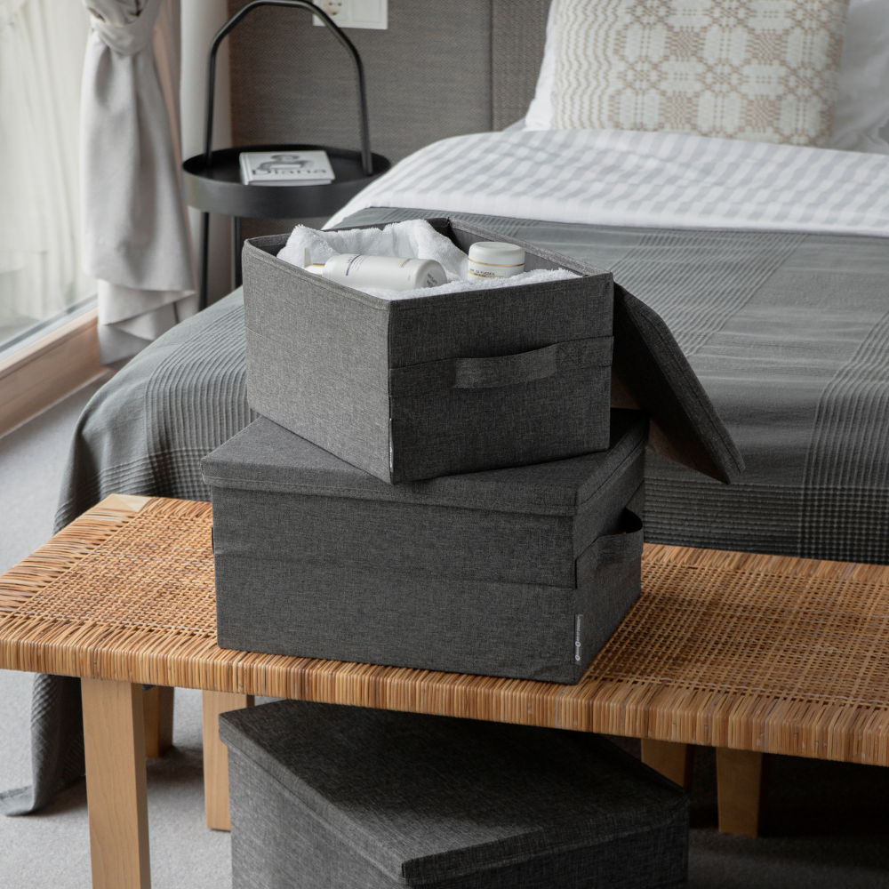 Twee Bigso SOFT opbergboxen in grijs, maat S (35x26x19 cm), waarvan er één open is en gevuld met toiletartikelen, staan op een gevlochten bankje aan het voeteneinde van een net opgemaakt bed met grijs en wit beddengoed in een moderne slaapkamer.