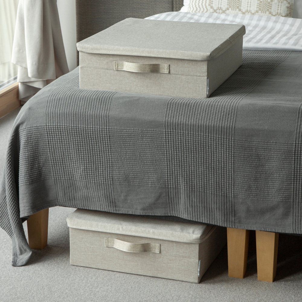 Twee beige SOFT-opbergboxen van Bigso, één op een grijs geruite dekbed en één die deels onder het bed met houten poten staat, in een gezellige, met tapijt beklede kamer.