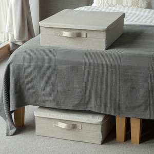 Twee beige SOFT-opbergboxen van Bigso, één op een grijs geruite dekbed en één die deels onder het bed met houten poten staat, in een gezellige, met tapijt beklede kamer.