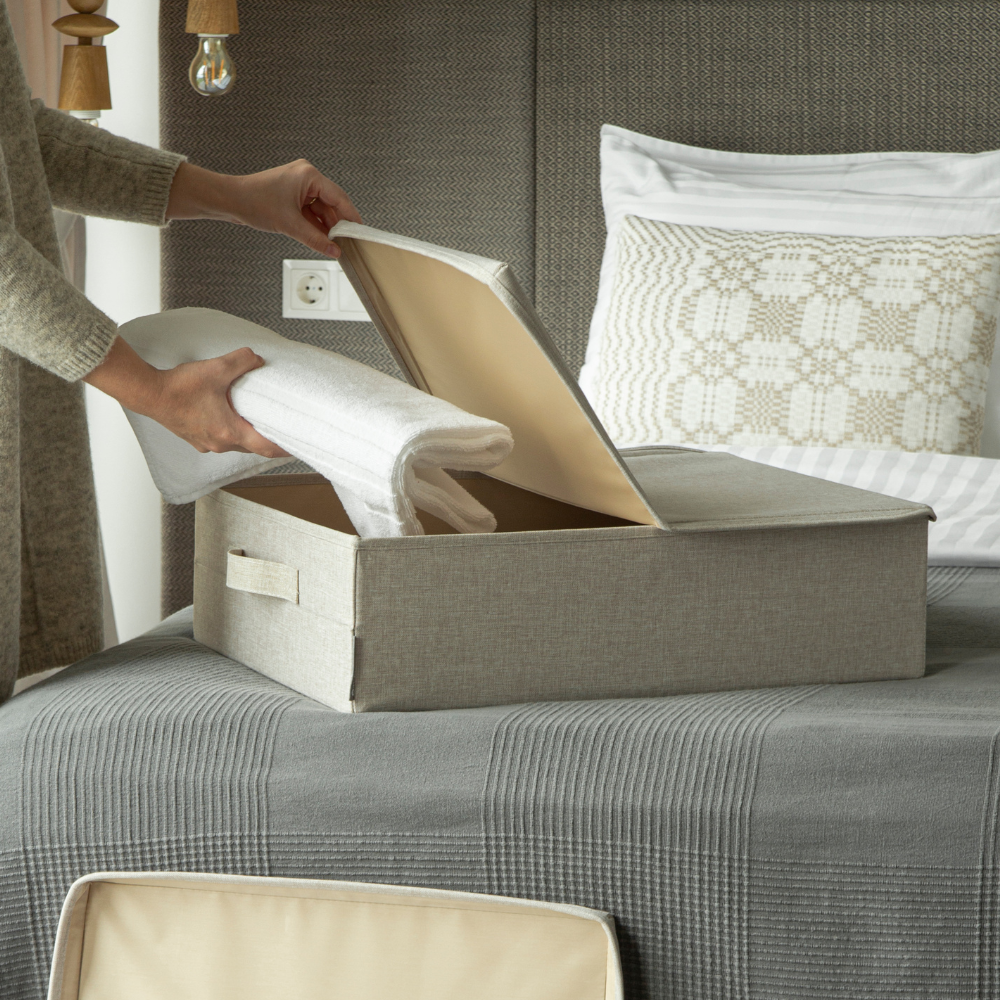 Iemand legt opgevouwen witte handdoeken in een beige SOFT-opbergbox van Bigso, bedoeld om onder het bed te bewaren, op een net opgemaakt bed met grijs beddengoed en een sierkussen, en creëert zo extra opbergruimte onder het bed.